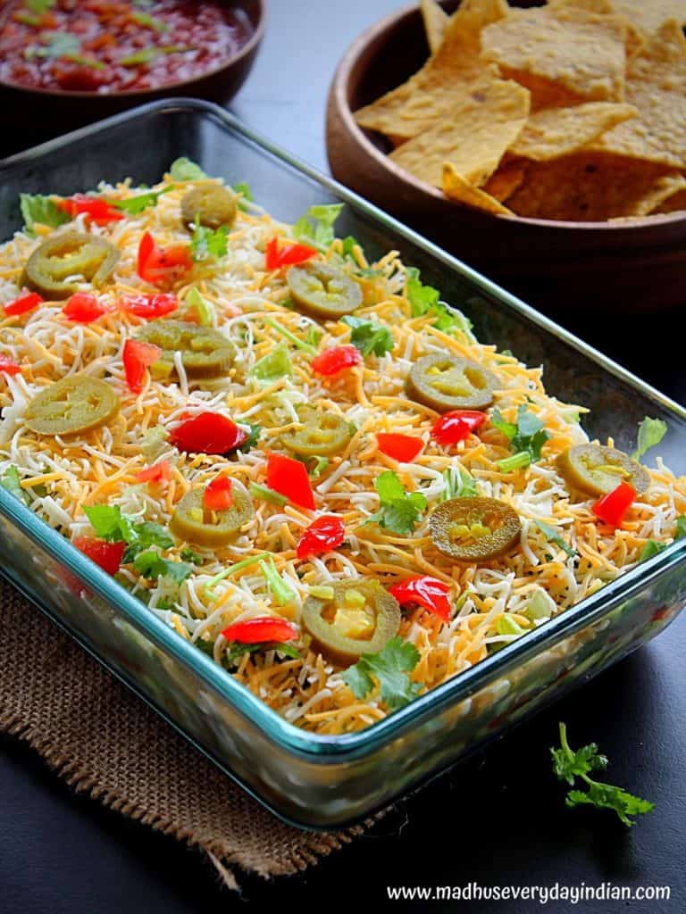 7 layer bean dip served in a glass tray garnished with jalapeno, tomato and coriander and served with tortilla chips and salsa