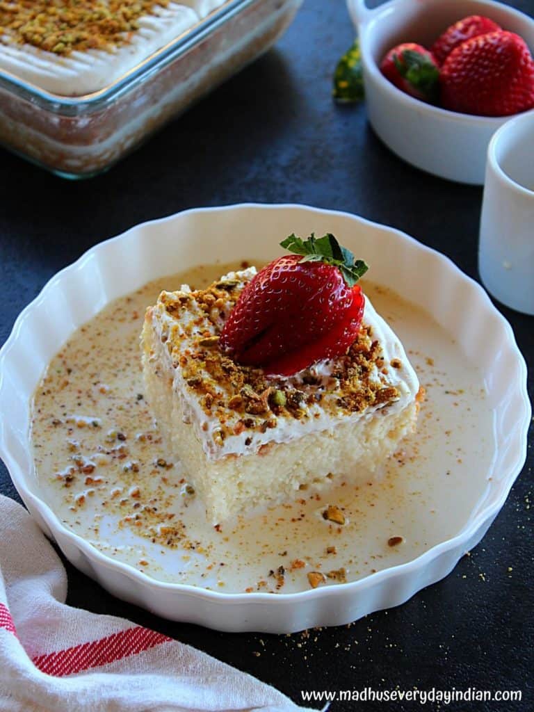 a slice of eggless tres leches cake served in a white plate garnished with 3 milk and pistachio and strawberry 