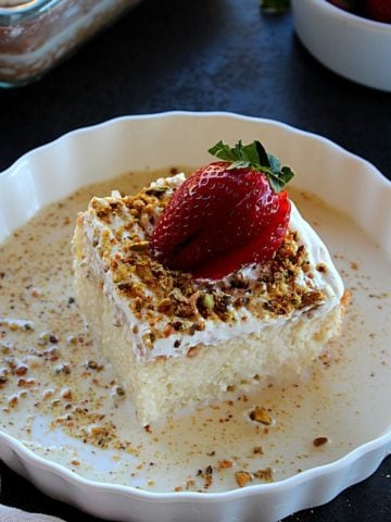 a slice of eggless tres leches cake served in a white plate garnished with 3 milk and pistachio and strawberry