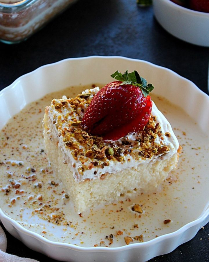 a slice of eggless tres leches cake served in a white plate garnished with 3 milk and pistachio and strawberry