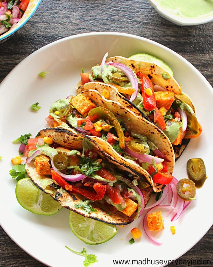 3 paneer tacos arranged in a white plate with lemon wedges